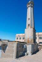 Historic scene at Alcatraz, San Francisco, USA. Weathered concrete structures of prison, lighthouse with rust, classical building, American flag on flagpole against clear blue sky. photo