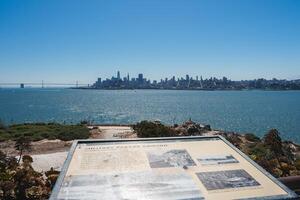 Scenic view of Alcatraz Island with MILITARY PARADE GROUND plaque, looking towards San Francisco skyline. Bay Bridge, boats visible. Sunny day, clear sky, no people. photo