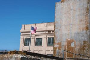 Alcatraz prison exterior in San Francisco, USA captured under a clear blue sky. Weathered American flag, aged facade, barred windows. A sense of abandonment and decay evident. photo