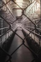 Interior view of Alcatraz prison in San Francisco, USA. Perspective through chain link fence shows cell blocks, barred doors, and stark lighting. Conveys isolation and historical atmosphere. photo
