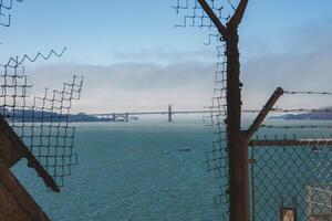 View through rusted bars and wire mesh inside Alcatraz overlooking San Francisco Bay. Golden Gate Bridge visible in distance with boats on calm water. photo