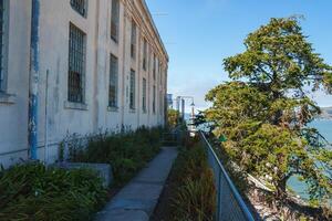 Alcatraz prison scene in San Francisco, USA. Aged building with barred windows, pathway with chain link fence, and greenery. Nature and history contrasted. photo