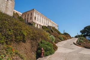 Exterior view of Alcatraz prison in San Francisco, USA. Large beige building atop hill with barred windows. Path, greenery, fire hydrant in foreground. Serene atmosphere with somber history. photo