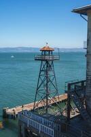 View from Alcatraz prison in San Francisco, USA. A guard tower stands on a pier with a weathered dome roof. A boat floats in the bay with a clear sky. photo