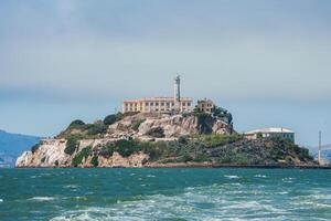 Alcatraz Island in San Francisco Bay, California, USA, viewed from the water. Shows rugged terrain, prison building, lighthouse, and choppy water under clear sky. photo
