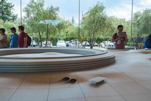 Interior of Apple Store by Apple office complex in Mountain View, CA. Features circular wooden table with product, person with iPad, bright, minimalist design, and lush outdoor view. photo