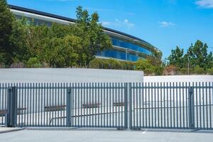 Modern architectural structure with curved design, glass panels, greenery and metal fence likely part of Apple office complex in Mountain View, California. photo