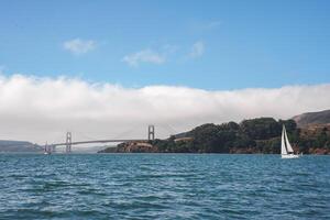 Serene view of Golden Gate Bridge over San Francisco Bay. White sailboat glides on calm water, hills covered in greenery, clear sky with scattered clouds. photo