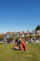Person in orange jacket sitting on grass near Painted Ladies in San Francisco. Sunny day, urban park setting with historic Victorian houses and blue sky. photo