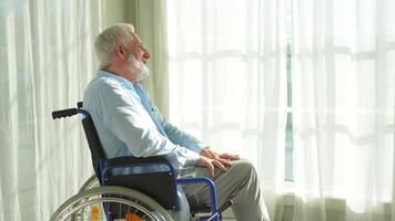 Sad senior man sitting in wheelchair looking out window in retirement home hospital. Old paralyzed man in chair for people with disability feel depressed. Lonely elderly sick man sitting on wheelchair video