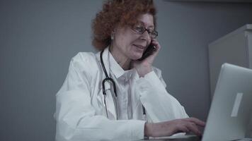 Middle-aged Caucasian female doctor sitting in her consulting room using phone and working on laptop. Elderly general practitioner talking on phone with patient while working on computer in clinic. video