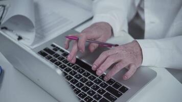 Senior woman doctor hands using laptop computer and working on checklist paper symptoms of patients.while sitting at desk in hospital office. She keeps all of her medical records on her laptop. video