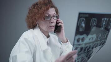 Elderly caucasian woman doctor analyze x-ray image and talk with mobile phone about diagnostics in medical office. Doctor specialist working with CT scan, talk to cellphone about radiography results. video