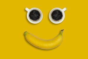 Happy morning concept. Two coffee cups and a banana shot on an bright yellow background. The composition of the objects looks like a smiley face. photo