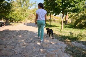 Woman walking a dog on a sunny day in a park pathway surrounded by lush green trees photo