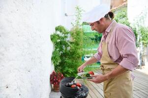 Man grilling vegetables on barbecue in outdoor setting with greenery and white wall background photo