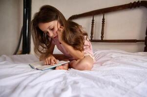 Cute smart little girl reading a book on her bed at home. Copy advertising space for text. The ideal image of a smart student pursuing a literary hobby. World Book Day concept photo