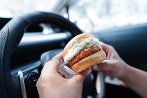 Asian woman driver hold and eat hamburger in car, dangerous and risk an accident. photo