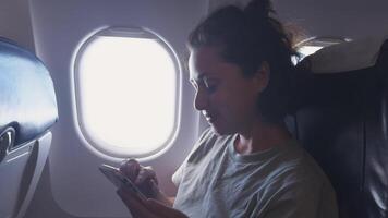 Passenger woman is flying in plane. Girl using phone sitting by airplane window. Traveling female with smartphone inside plane enjoying flight. Woman using wifi connection on board. Traveling girl video