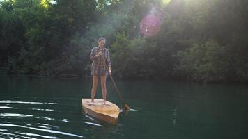 Woman is standing and floating on a small raft in the middle of the river. She is using an oar to row. video