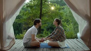 Young couple sits on the terrace, in front of a tent in a forest camping area, surrounded by trees. They hold hands and exchange sweet kisses. video