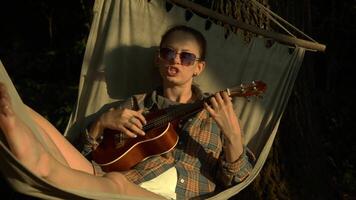 A young woman is playing the ukulele and singing while lying in a hammock, enjoying the sunset. video