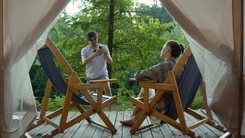 Man takes a photo of his girlfriend sitting in an armchair, in front of a tent, at the campsite where they are staying. video