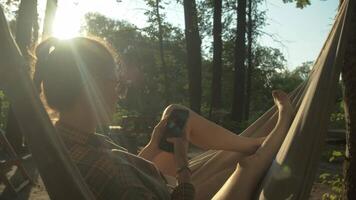 A girl is sitting in a hammock, swinging in nature, while she is using social media on her phone. video