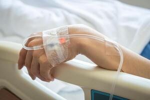 Close up of patient woman hand with receiving intravenous fluid directly into a vein while her hand touching bed rails on hospital bed. Conceptual of patient woman resting in hospital. photo