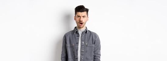 Shocked and offended man frowning, looking at camera with opened mouth, feeling hurt by cruel words, standing disappointed on white background photo