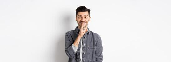 Happy guy shushing at camera, smiling with finger on lips, asking to keep quiet, telling big secret or making surprise, standing on white background photo