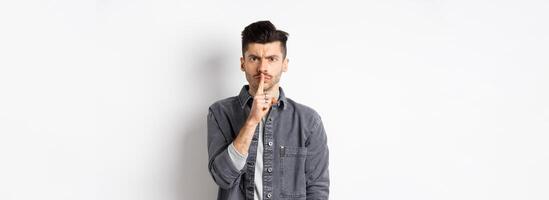 Be quiet. Angry man frowning and shushing, telling to calm down, press finger to lips as scolding bad behaviour, standing on white background photo