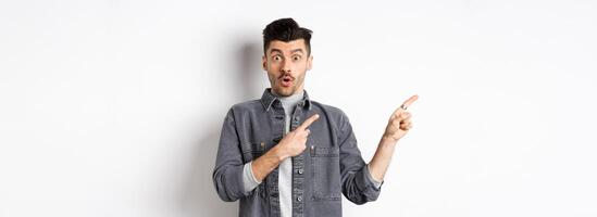 Excited man pointing left at logo and look with disbelief, saying wow at camera, checking out cool promo discount, standing on white background photo