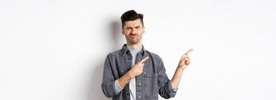 Skeptical and disappointed young man pointing fingers left, grimacing and shaking head in rejection, dislike bad product, standing on white background photo