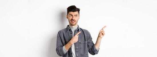 Skeptical and doubtful young man pointing fingers left at logo, look with dislike and confusion, standing on white background photo