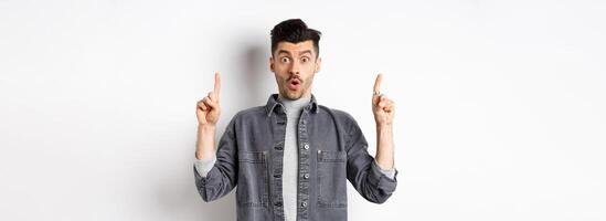 Excited man checking out promo offer, gasping amazed and pointing fingers up, making cool announcement, standing on white background photo