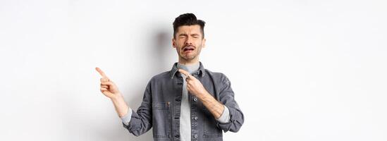 Crying man whining and complaining, pointing fingers right at sad news, standing miserable on white background photo