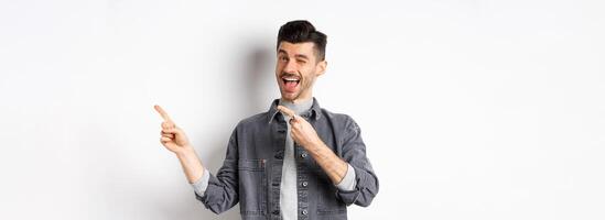 Handsome smiling guy making announcement, winking and pointing at upper right corner, showing logo, inviting to check out advertisement, standing on white background photo