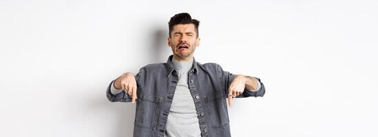 Sad crying man pointing fingers down, sobbing miserable, showing upsetting news, standing in denim jacket on white background photo