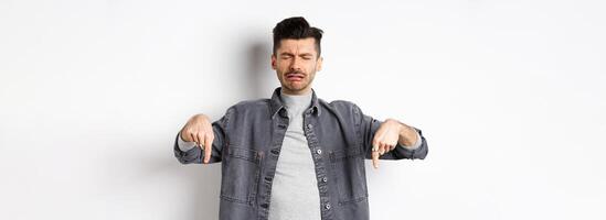Sad crying man whining, pointing fingers down disappointed, showing bad news, standing upset on white background photo