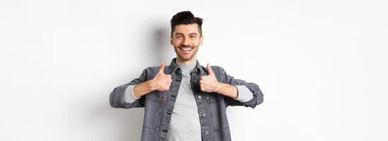Handsome smiling man showing thumbs-up in approval, praise good work, satisfied with nice choice, recommending something cool, standing on white background photo