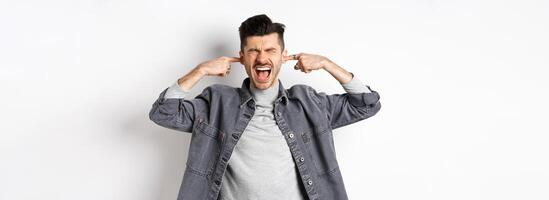 Annoyed guy shut ears and screaming, displeased by loud noise, yelling to stop noisy music, standing on white background photo