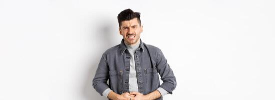 Man with stomach ache frowning and touching belly, having food poison, suffer diarrhea, standing on white background photo
