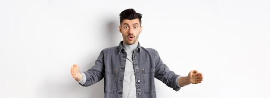Excited caucasian man showing big size, shaping large object and look amazed, standing on white background photo