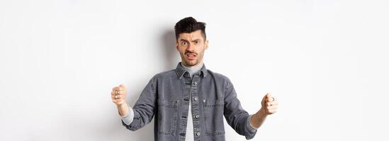 Skeptical young man showing big size object and frowning doubtful, standing reluctant on white background photo