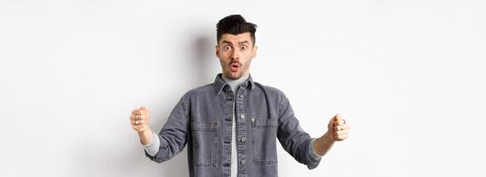 Confused man holding big size object and looking shocked, shaping large thing, standing on white background photo
