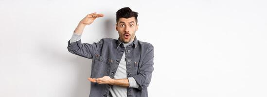 Cheerful guy smiling and showing big size, showing large thing and looking excited, standing against white background photo
