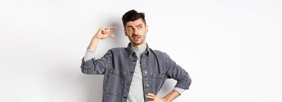 Pensive young man looking aside and thinking, showing small size object, pondering something little or tiny, standing on white background photo