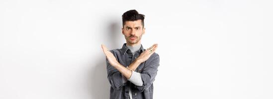 Angry man show cross gesture to stop or say no, frowning and looking serious, disagree and prohibit bad situation, standing on white background photo
