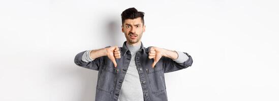 Disappointed guy showing thumbs down and frowning, looking skeptical at something bad, dislike and disapprove awful thing, standing on white background photo